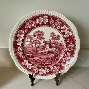 May include: A round ceramic plate with a scalloped edge featuring a deep red transferware pattern. The center depicts a landscape with a stone bridge, a castle, and trees. The border is decorated with floral motifs and small circular patterns.