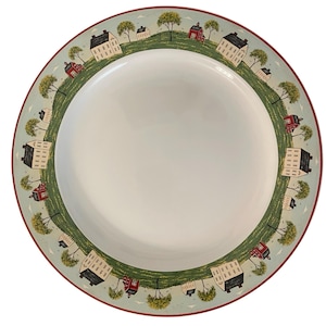 May include: A white ceramic plate with a decorative border featuring a village scene. The border includes small houses, trees, and a green landscape against a light blue sky. The rim is red.