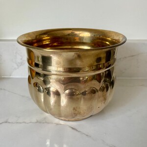 May include: A polished brass planter featuring a wide, flared rim and a rounded body with a series of vertical, scalloped indentations around the lower section. The metal surface has a reflective, warm gold tone with subtle variations in color.