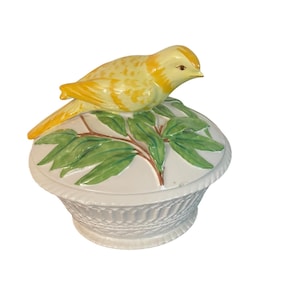 May include: A decorative ceramic trinket box with a yellow bird perched on top. The lid features green leaves and a white base, while the basket-like bottom is white. The bird is yellow with orange accents.
