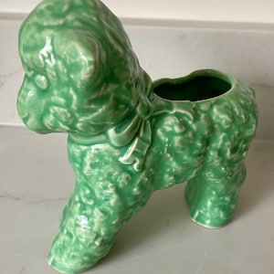 May include: A glossy, light green ceramic planter shaped like a lamb. The lamb figure features a textured, curly wool surface, a smooth bow tied around its neck, and an open cavity on its back for holding small items or plants.