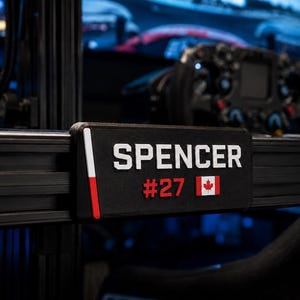 May include: A black rectangular nameplate attached to a racing simulator frame. The plate features the white text SPENCER, the red number #27, a small Canadian flag, and a vertical white and red stripe on the left side against a black background.