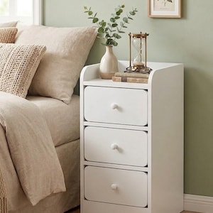 May include: A white bedside table with three drawers and a curved top. A vase with greenery, an hourglass, and books sit on top. A framed botanical print hangs on the wall. The bed has beige bedding and pillows.