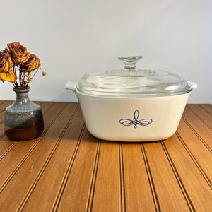 May include: A white rectangular ceramic casserole dish with a clear glass lid and a black decorative flourish design on the front, accompanied by a small, two-toned blue and brown ceramic vase containing three dried orange roses.