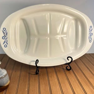 May include: An oval white ceramic platter featuring a central grooved design for drainage and blue floral accents on both sides. The platter is supported by a black metal stand, and a small blue and brown ceramic vase with dried flowers sits nearby.