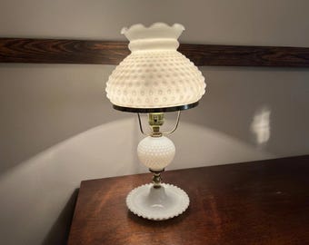 May include: A white hobnail glass table lamp with a ruffled shade. The lamp has a round base and a spherical body, with brass-coloured metal accents. The shade is a classic shape, and the lamp is lit.
