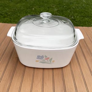 May include: White casserole dish with a clear glass lid. The dish has a floral design in pastel colors on the front. The dish has two white handles on either side. The lid has a clear knob handle.
