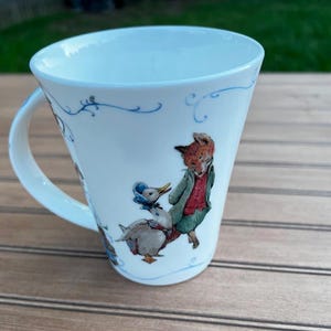 May include: White ceramic mug with a curved handle, featuring a whimsical illustration of a fox and a duck. The mug has a light blue rim and decorative blue floral accents. The fox is wearing a green jacket and the duck is wearing a blue bonnet.