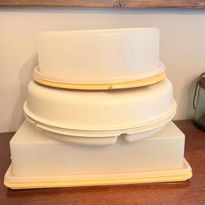 Puede incluir: Una pila de recipientes de almacenamiento de alimentos de plástico blanco roto vintage con detalles amarillos. Los recipientes tienen varias formas y tamaños, incluyendo una base rectangular y tapas redondas. Las tapas tienen un borde amarillo. Apilados sobre una superficie de madera.