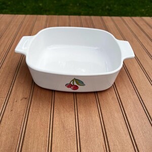 May include: White rectangular baking dish with two handles. The dish features a cherry design on the front. The dish is sitting on a wooden surface.