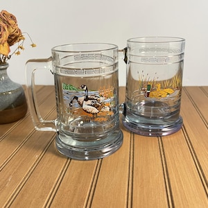 May include: Two clear glass beer mugs featuring a Greek key pattern around the top rim. One mug displays a graphic of two Canada geese in a marsh, while the other mug shows a mallard duck swimming in reeds. Both mugs have sturdy handles.