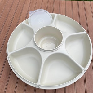 Op de afbeelding: Een witte plastic serveerschaal met een centrale dipschaal en zes omliggende compartimenten. Een bijpassend deksel rust op de schaal. De schaal is ontworpen voor het serveren van snacks of hapjes. De schaal staat op een houten oppervlak.