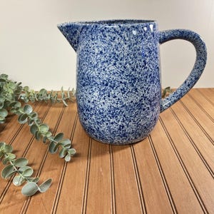 May include: A ceramic pitcher featuring a speckled pattern of dark blue and white. The vessel has a rounded body, a curved handle on the right side, and a pointed pouring spout at the top rim. The surface texture appears uniform and consistent.