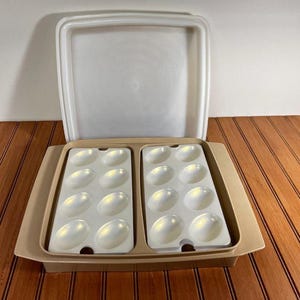 May include: A beige plastic egg storage container with a matching lid. The base features two removable white inserts, each containing eight oval-shaped indentations designed to hold sixteen eggs in total. The lid is upright and detached.