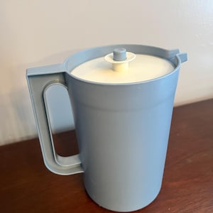 May include: A light blue plastic pitcher with a matching handle and lid. The pitcher has a cylindrical shape and a white lid with a small, light blue knob. The pitcher is sitting on a dark brown surface.