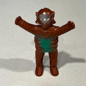 May include: A brown action figure with a silver face shield and outstretched arms. The figure has a green midsection and a textured, scaly appearance. The toy is standing on a light grey surface.