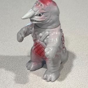 May include: A grey and red monster figurine with a long beak and clawed feet. The figure has a red patch on its head and chest. The toy is standing on a light grey surface.