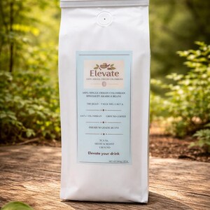 Op de afbeelding: Een witte zak koffie van het merk Elevate, met een lichtblauw etiket. Het etiket luidt "100% Single Origin Colombian Specialty Arabica Beans". De zak staat op een houten oppervlak, met een vage groene achtergrond. 140g.