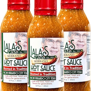 May include: Three bottles of Lala's Cafe Serrano Hot Sauce. Each bottle has a red cap and a label with a portrait of a man wearing glasses, a red chili pepper, and the text "Hot Sauce". The bottles are 12 fl oz / 355ml.