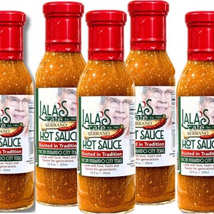 May include: Five bottles of Lala's Cafe Serrano Hot Sauce with red caps and labels featuring a portrait and the text "Hot Sauce." The labels also state "Rooted in Tradition" and "From Mirando City Texas." Each bottle contains 12 fl oz.