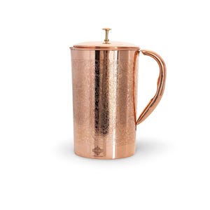 May include: A cylindrical copper pitcher featuring an intricate floral and paisley pattern etched across its surface. The pitcher includes a matching lid with a small knob handle, a curved side handle, and a smooth base with a small logo.