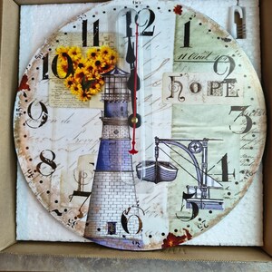 May include: A round wall clock featuring a lighthouse illustration, a small boat, and a cluster of yellow flowers. The clock face displays black numbers, cursive script, and the word HOPE. It has black hour and minute hands with a red second hand.