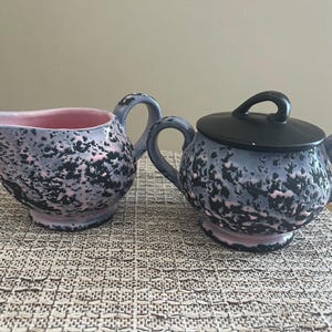 May include: A ceramic creamer and a lidded sugar bowl featuring a textured, mottled glaze in shades of black, light blue, and soft pink. The creamer has a curved handle and spout, while the sugar bowl includes two side handles and a black lid.