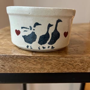 May include: A cream-coloured ceramic ramekin featuring a hand-painted design of three dark blue geese walking in a line, flanked by two small red hearts on either side. The bowl has a smooth, slightly speckled finish and a simple, cylindrical shape.