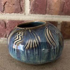 May include: A round, ceramic vase with a glossy finish. The vase is primarily blue with streaks of tan and brown, and features a decorative pattern. The interior is a darker blue. The vase is sitting on a gray surface.