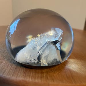 May include: A clear, spherical glass paperweight containing a jagged, white, crystalline mineral formation at its center. The smooth, rounded exterior of the glass sphere reflects light, while the internal structure appears textured and opaque.