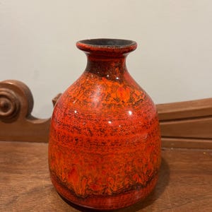 May include: A ceramic vase with a bulbous body and a narrow neck. The vase is primarily orange with dark brown and black marbling. The interior of the neck is black. The vase is a decorative item.