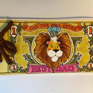 May include: A rectangular card with a lion illustration, a crown, and the words "ONE-TIME FREE OFFER" and "BEST DAD". The card has a brown, braided attachment on the left side. The lion has a yellow face and a brown mane.