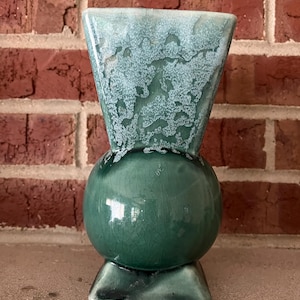Könnte beinhalten: Eine türkisfarbene Keramikvase mit kugelförmigem Sockel und einem ausgestellten, strukturierten Oberteil. Die Vase hat eine glänzende Oberfläche und steht auf einem dreieckigen Sockel. Der Hintergrund zeigt eine rote Backsteinmauer.