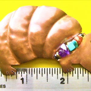 May include: A copper-colored armadillo figurine with a beaded collar of purple, turquoise, and red stones. The armadillo is approximately 3 inches long, with a textured surface and detailed feet. The background is yellow.