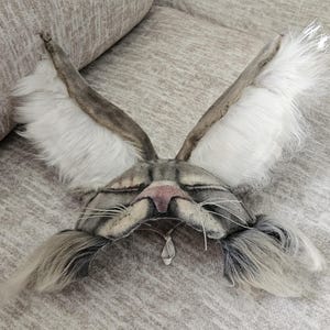 May include: A handmade gray and white cat mask with long, upright ears and a pink nose. The mask features long, gray whiskers and a small, dangling pendant. The ears are lined with white fur.