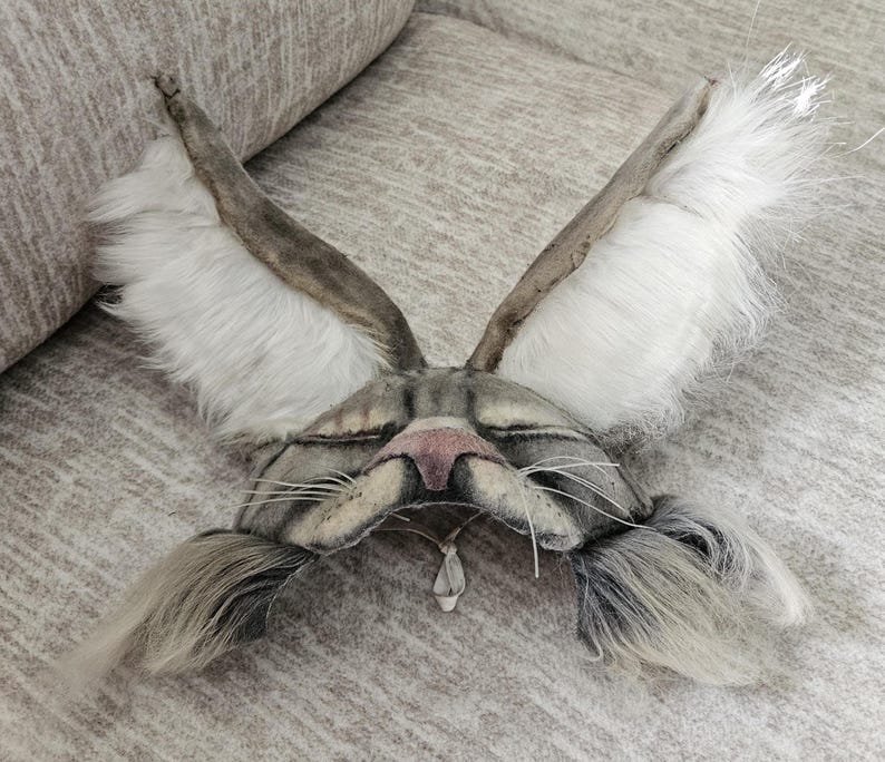 May include: A handmade gray and white cat mask with long, upright ears and a pink nose. The mask features long, gray whiskers and a small, dangling pendant. The ears are lined with white fur.