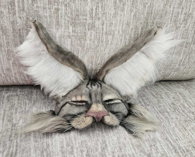 May include: A gray and white rabbit mask with long, upright ears and a pink nose. The mask has closed eyes and long, gray whiskers. The ears are lined with white fur. The mask is made of a soft, textured material.