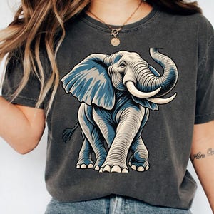 May include: A charcoal gray short-sleeve t-shirt featuring a large graphic illustration of an elephant in shades of blue and cream. The elephant is depicted in a forward-facing stance with its trunk raised and tusks visible, centered on the chest.