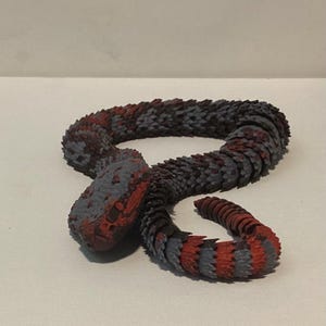 May include: A flexible, articulated snake figurine featuring a segmented body with a repeating pattern of matte red and dark gray scales. The snake is coiled in a resting position, showcasing a textured, layered design from its head to its tail.