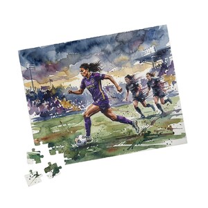 May include: A watercolour puzzle depicting a soccer match. A player in a purple jersey with "SPIRIT" on it is running with the ball, followed by two teammates. The background shows a stadium and a cloudy sky. Includes a few detached puzzle pieces.