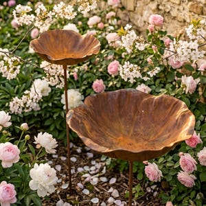 May include: Two weathered, copper-colored bird baths with a leaf-like design stand in a garden. The bird baths are surrounded by pink and white flowers, with a stone wall in the background. The garden is filled with blossoms.