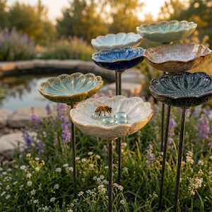 May include: A collection of ceramic bird baths in various colors, including blue, green, and cream, with a flower-like design. Each bath is mounted on a dark metal stem. A bee is visible on one of the baths, along with several clear glass beads.