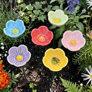 May include: A collection of colorful ceramic flower garden stakes. The stakes feature flower heads in blue, red, pink, yellow, purple, and green, each with a yellow center. A bee rests on the yellow flower. The stakes are in a garden setting.