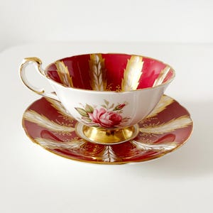 May include: A fine bone china teacup and saucer set. The teacup features a red interior with gold detailing and a floral motif. The saucer is red with gold patterns. The set is a classic design.