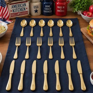 Puede incluir: Un conjunto de cubiertos dorados, que incluye tenedores, cucharas y cuchillos, dispuestos sobre un mantel azul oscuro. La escena está ambientada con una decoración de cocina de temática americana.