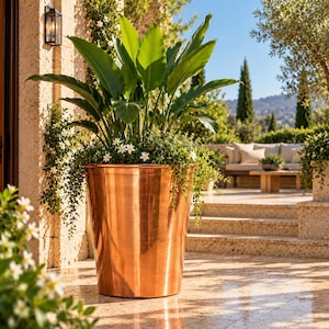 Puede incluir: Una maceta cilíndrica grande de cobre pulido que contiene una planta verde vibrante con hojas anchas y erguidas y pequeñas flores blancas que caen sobre el borde, situada en un patio de piedra en un entorno exterior soleado.