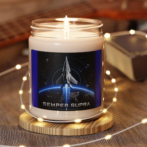 May include: A clear glass jar candle with a white wax fill and a lit wick. The label features a graphic of a space shuttle launching above the Earth with the text "SEMPER SUPRA" printed in white capital letters at the bottom of the label.