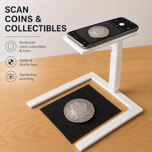 May include: A white smartphone stand with a coin on a black cloth. The phone is positioned above the coin, with the text "SCAN COINS & COLLECTIBLES" in the background. The stand is designed for hands-free scanning.