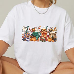 Pode incluir: Uma t-shirt branca de manga curta com uma impressão gráfica colorida de quatro copos de bebidas decorados com elementos de outono e fantasia, incluindo abóboras, livros, um chapéu de bruxa, um cachecol e uma pequena coruja branca com uma carta.