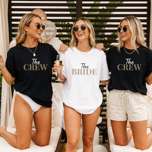 Op de afbeelding: Twee zwarte t-shirts met de tekst The CREW in goud en één wit t-shirt met de tekst The BRIDE in goud. De t-shirts hebben korte mouwen en een ronde hals. De kledingstukken worden gedragen door drie personen die champagneglazen vasthouden.